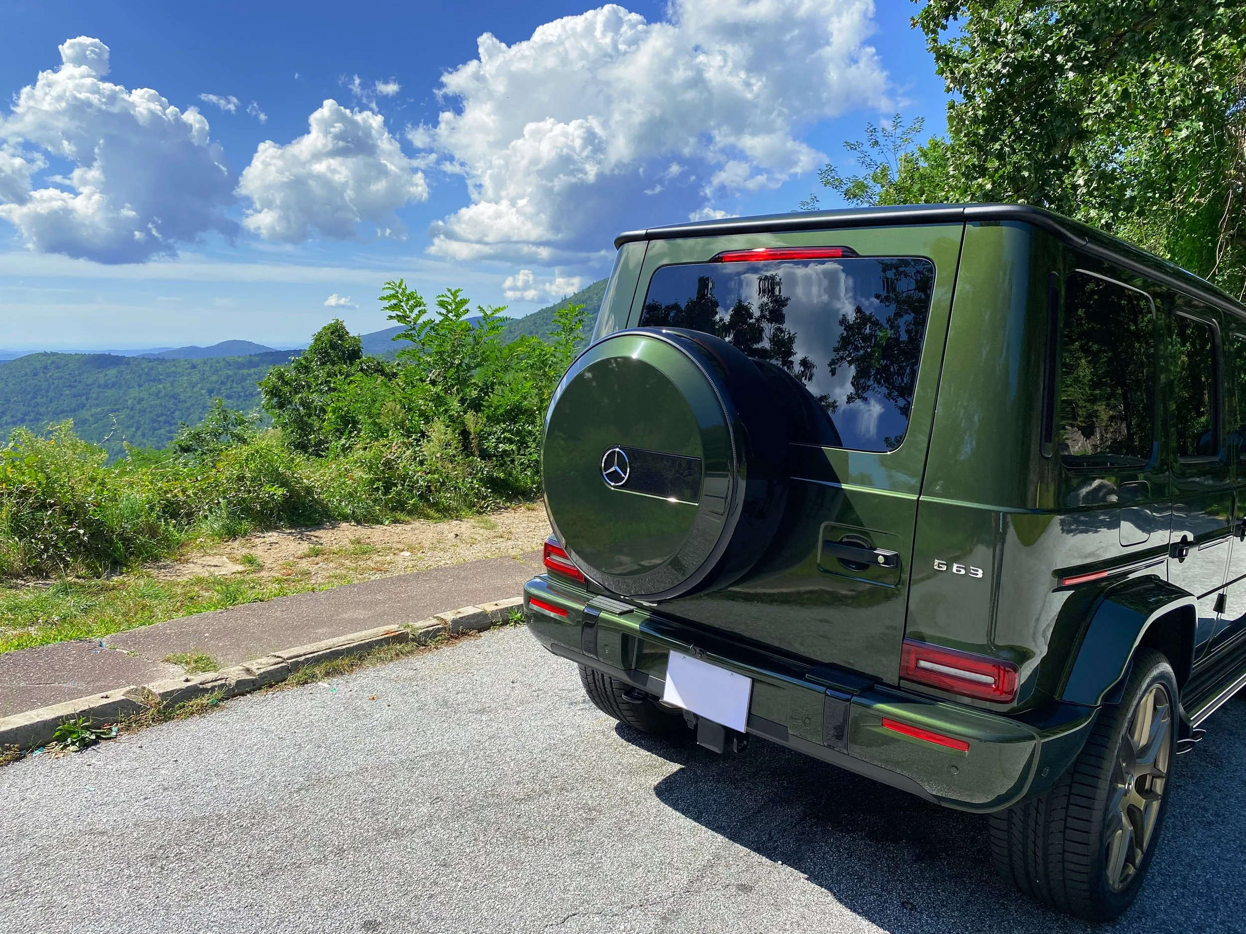 Rear view of an olive green Mercedes-Benz G-Class SUV parked on a mountain overlook with a scenic view of lush green mountains and a partly cloudy sky