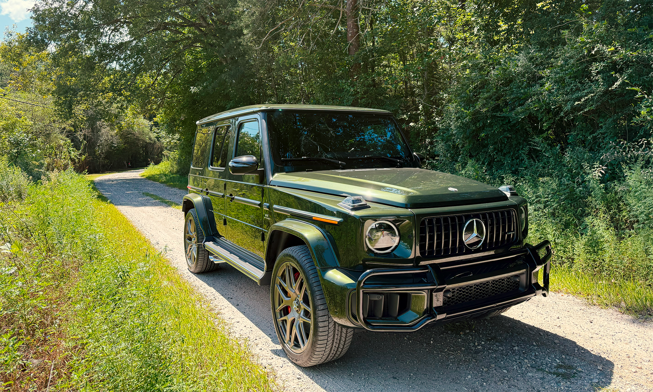 An olive green Mercedes-Benz G-Class SUV parked on a dirt road surrounded by green trees and foliage.