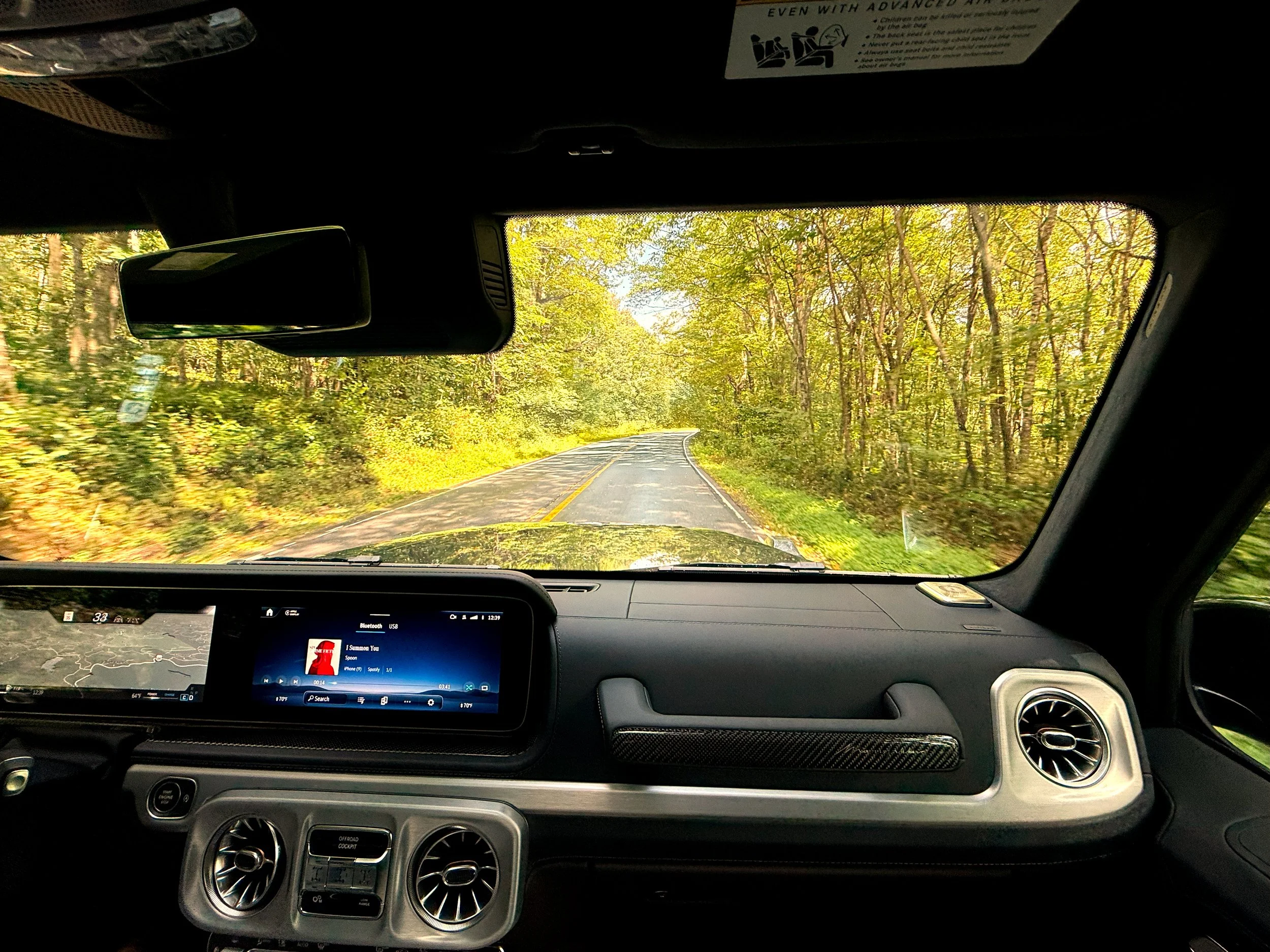 Inside a 2025 Mercedes-Benz G63 vehicle, looking out at a winding road through a forest with green trees.