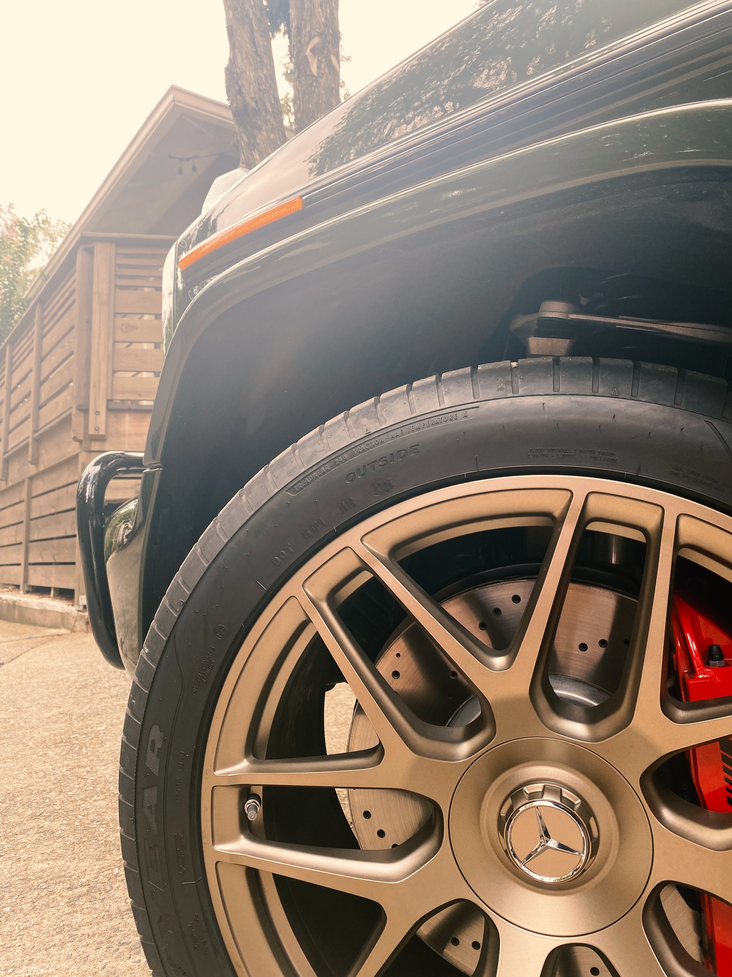 Close-up of a 2025 Mercedes-Benz G63 car wheel with a gold alloy rim, red brake caliper, and disc brake, parked near a wooden fence and trees.