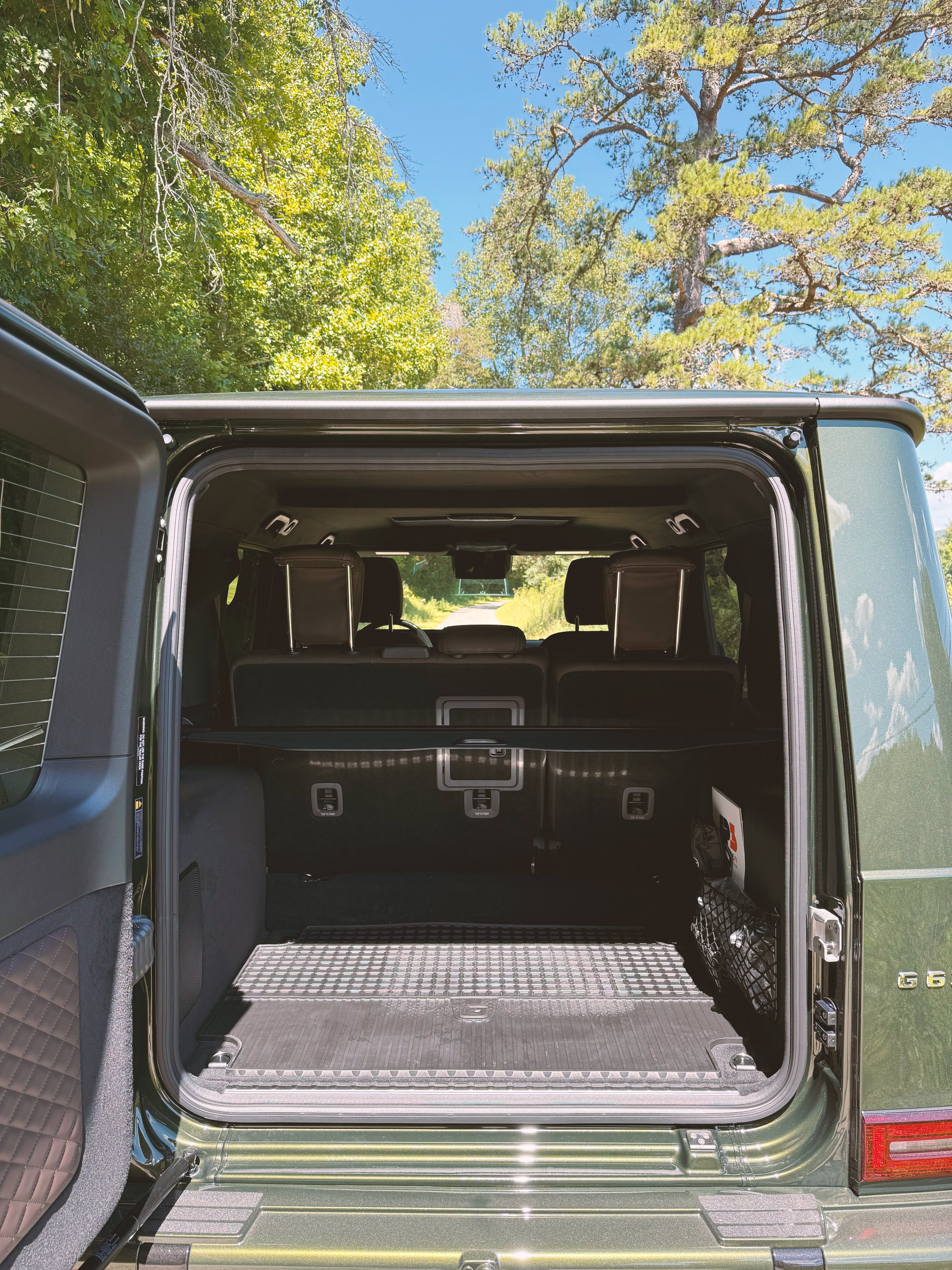 Open trunk of an olive green 2025 Mercedes-Benz G63 SUV parked outdoors with trees and blue sky in the background.