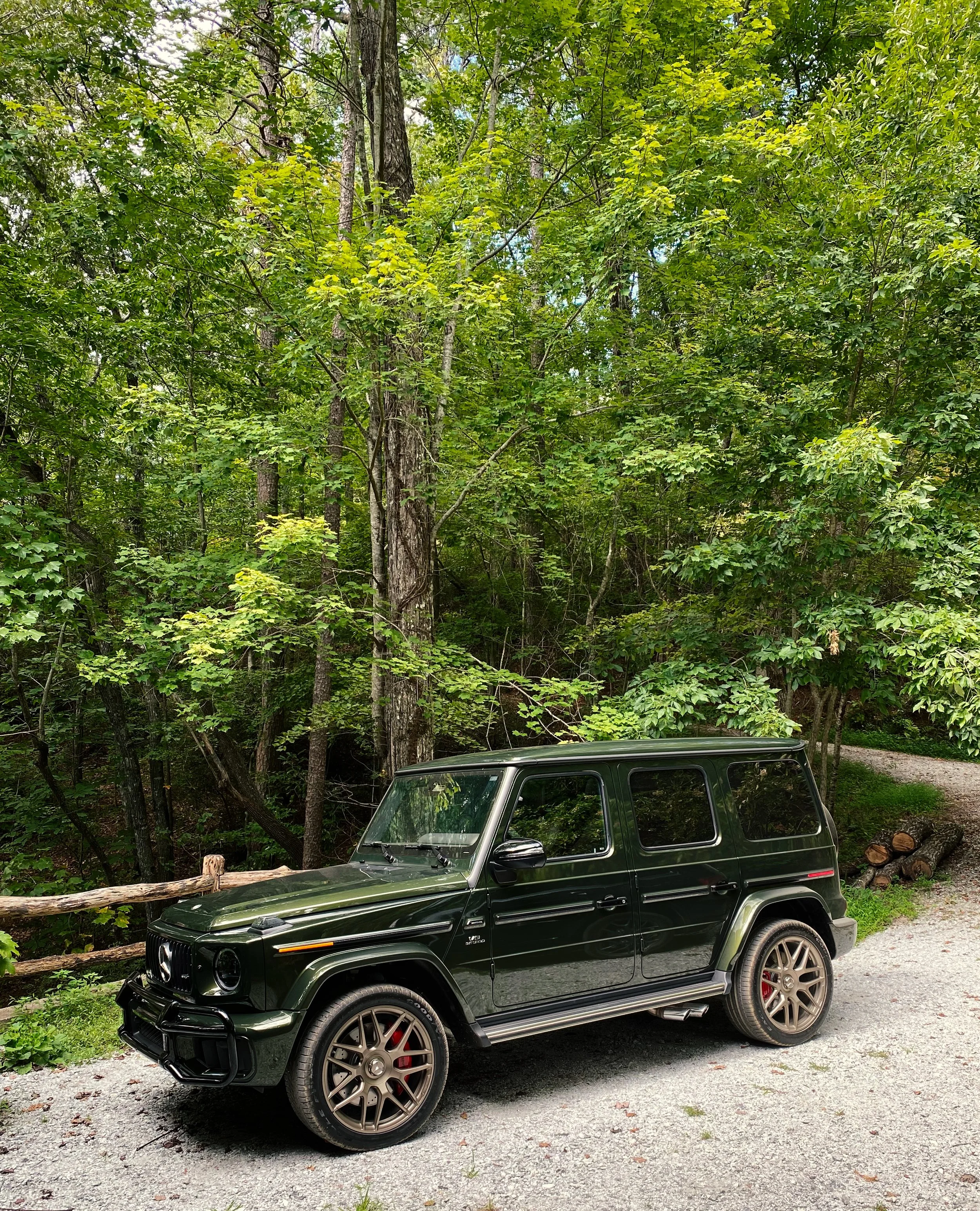 An olive green 2025 Mercedes-Benz G63 SUV parked on a gravel road in a wooded area with green trees and logs in the background.