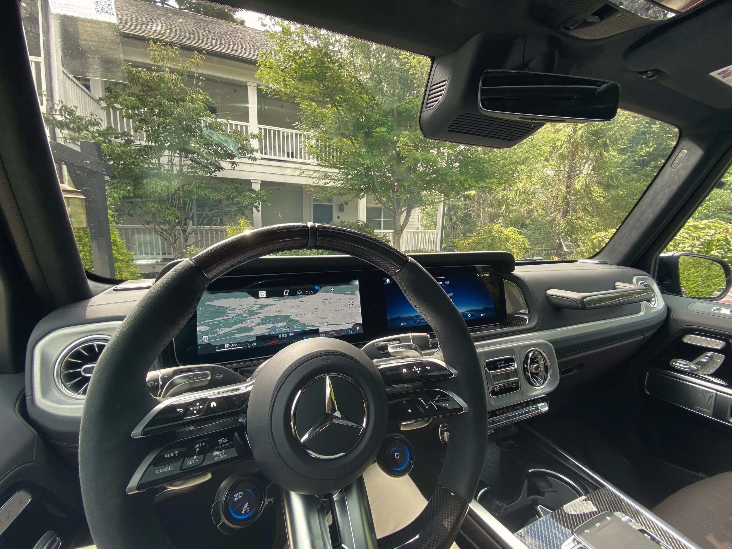 The interior of a luxury 2025 Mercedes-Benz G63 vehicle showing the dashboard, steering wheel, and digital display screens. Outside, a residential house with a porch and trees is visible through the windshield.