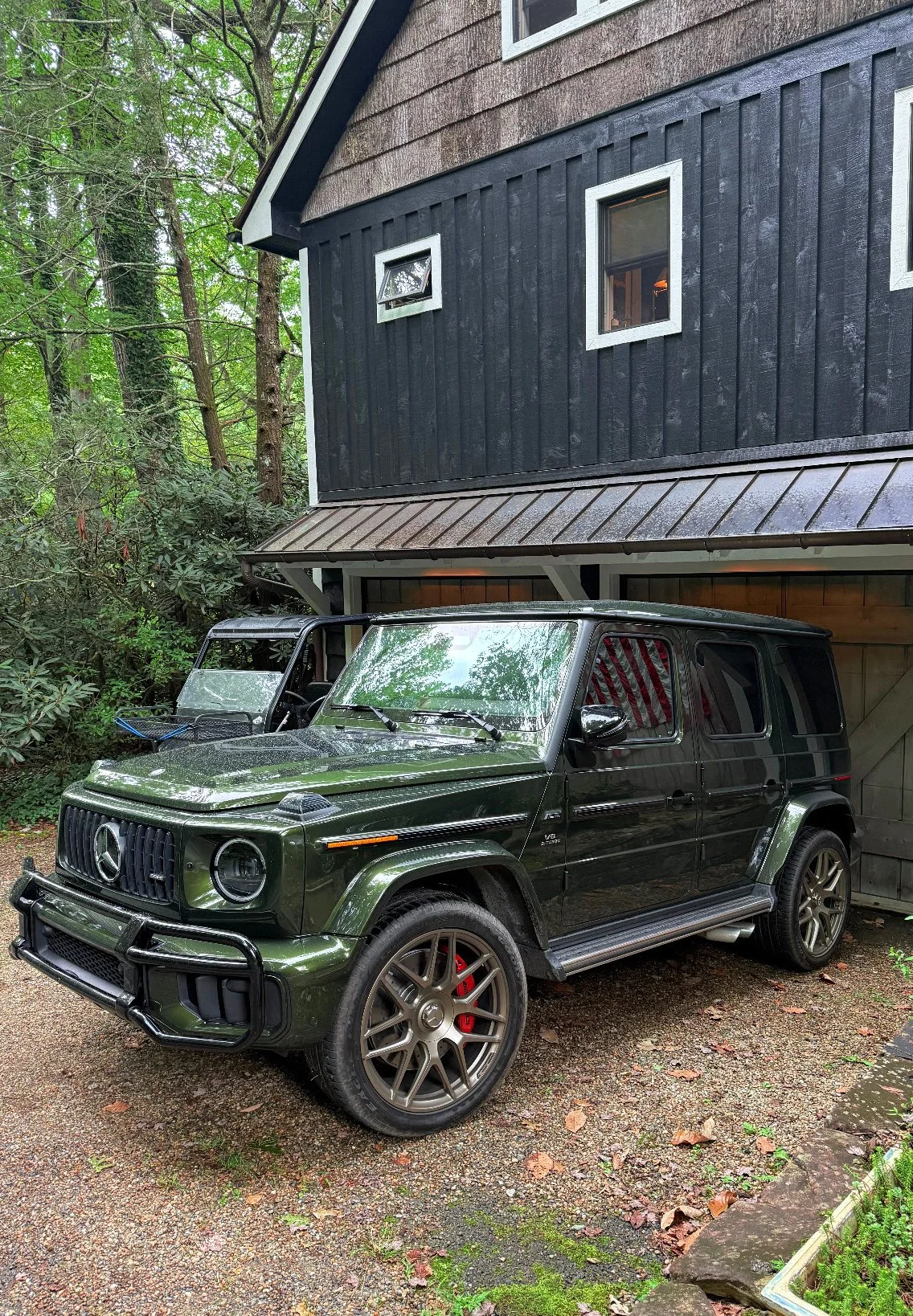 An olive green Mercedes-Benz G-Class SUV parked on a gravel driveway next to a black wooden house with small windows.