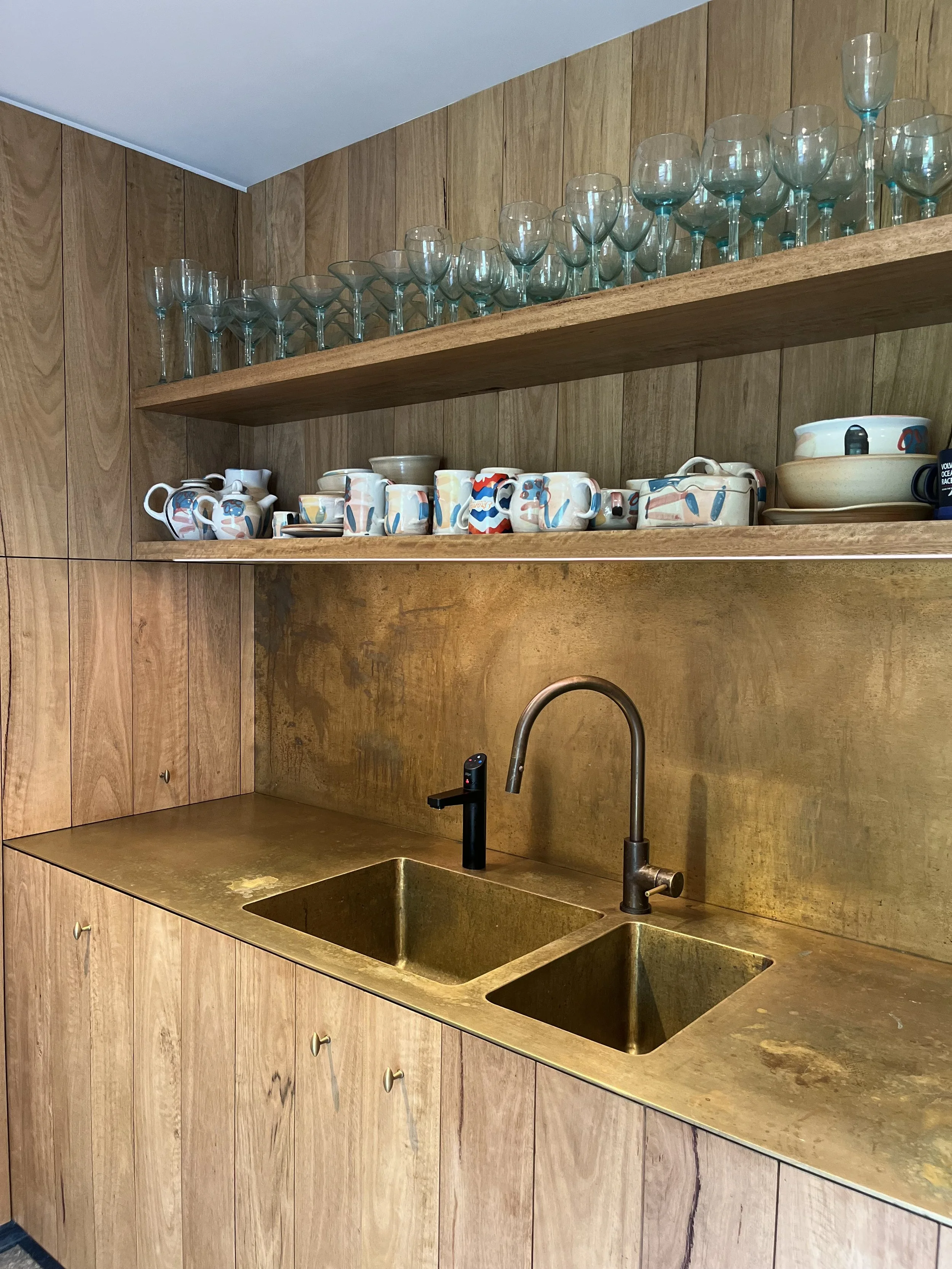 A kitchen with wooden cabinets, a gold-toned countertop and backsplash, and a gold-toned sink. An open wooden shelf above holds various glassware including wine glasses and cups with colorful patterns.