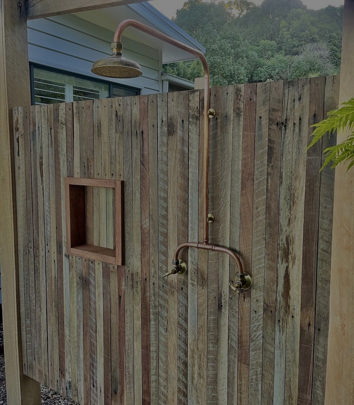 An outdoor shower with a copper pipe and showerhead mounted on a weathered wooden privacy wall, with a small open cubby built into the wall.