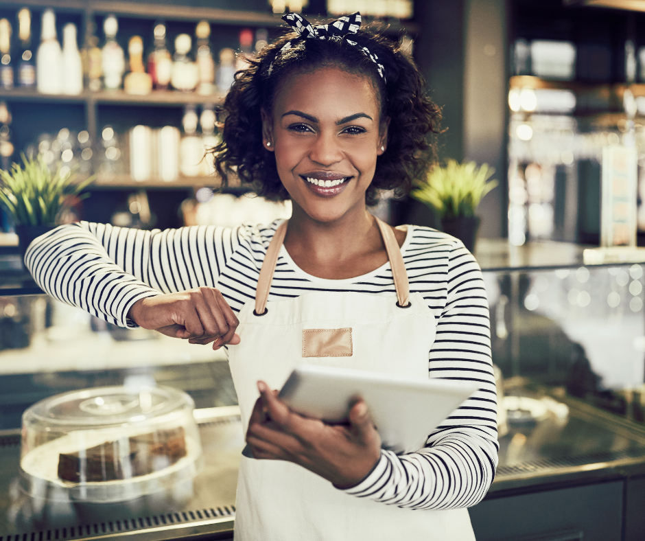 Smiling woman in a cafe wearing a striped shirt and an apron, holding a tablet.