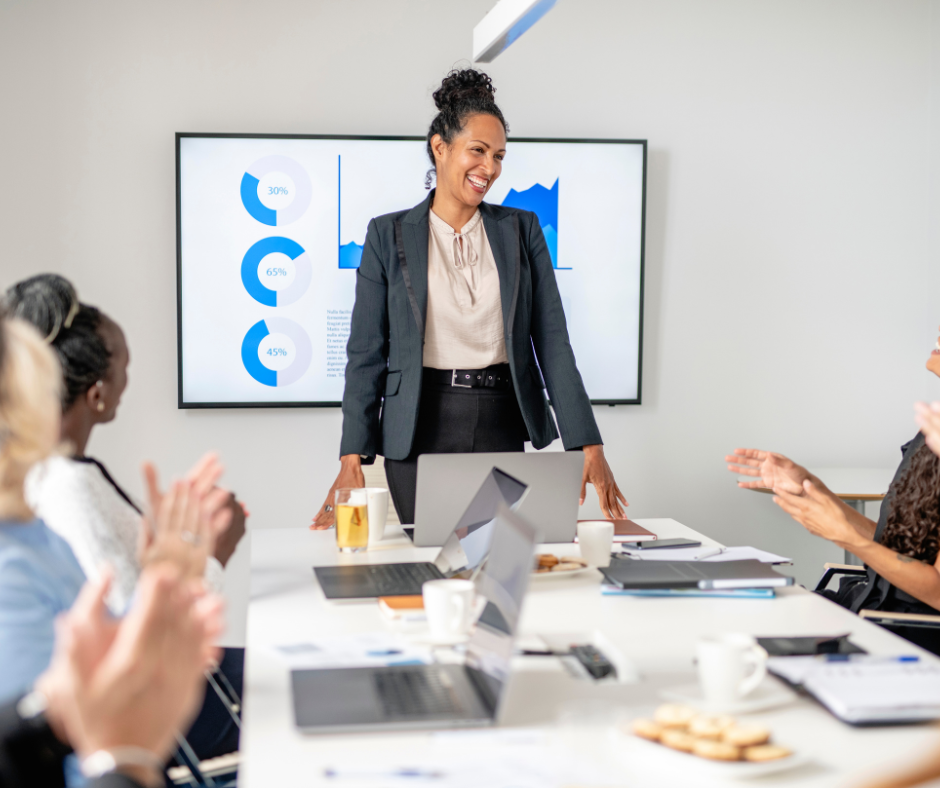 Businesswoman leading a meeting with colleagues, smiling and standing at a conference table with laptops, papers, and coffee cups, in front of a presentation screen with charts.