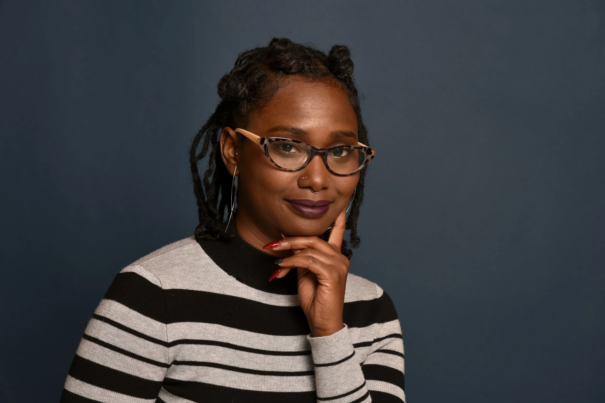 A woman with dark skin, curly hair in small buns, wearing glasses with a turtle shell pattern, a nose ring, and a striped sweater, smiling and holding her chin with her hand against a dark background.
