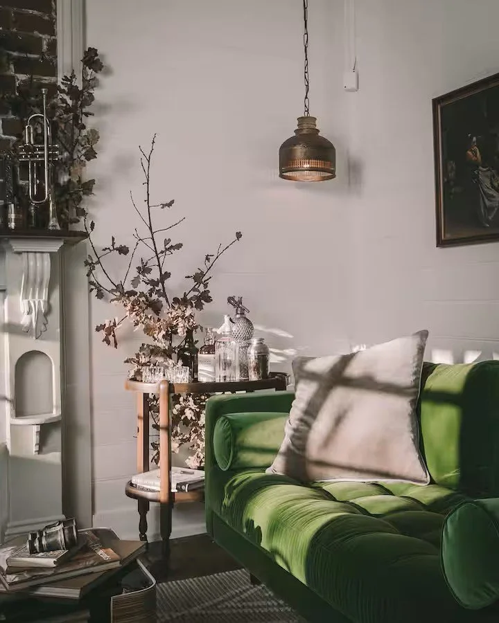 A cozy living room corner with a green velvet sofa, a beige pillow, a small side table with decorative bottles, a large branch with pink blossoms in a vase, and a wall-mounted pendant light casting shadows.