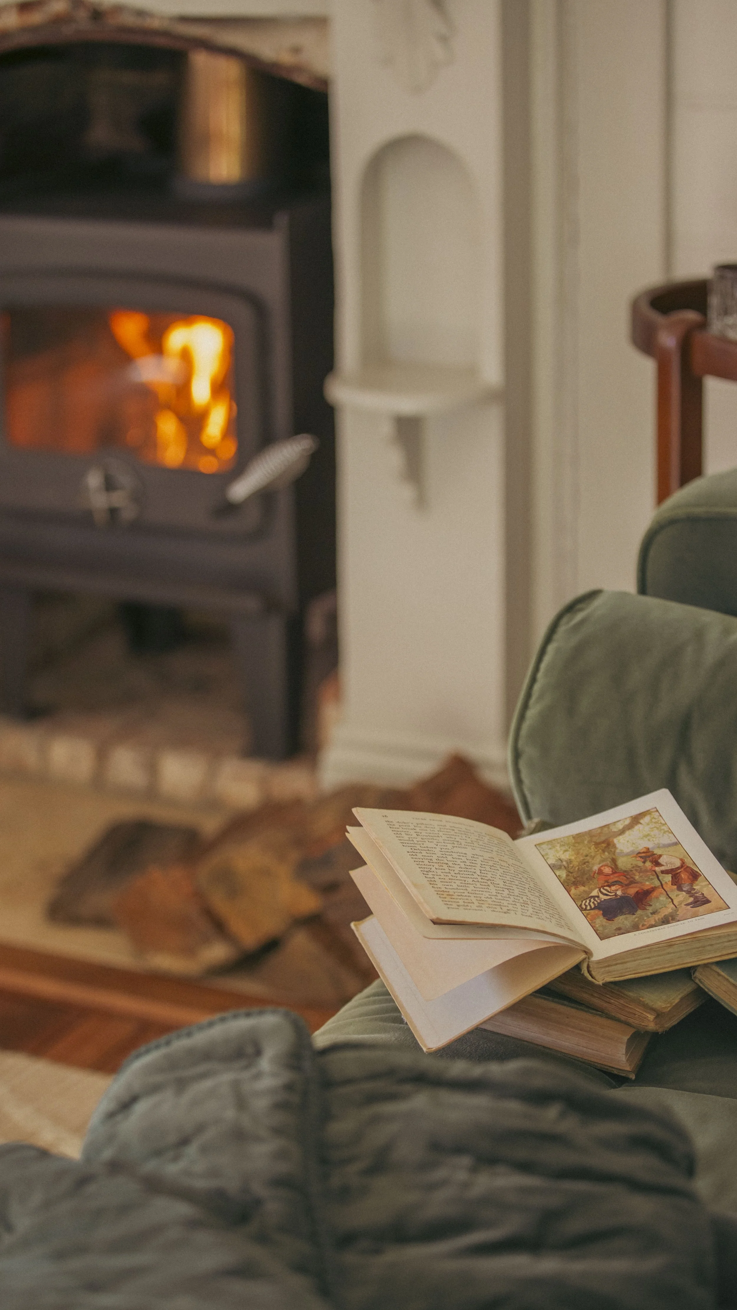 Open book with colorful illustrations resting on a cushioned chair in front of a cozy fireplace with a crackling fire.