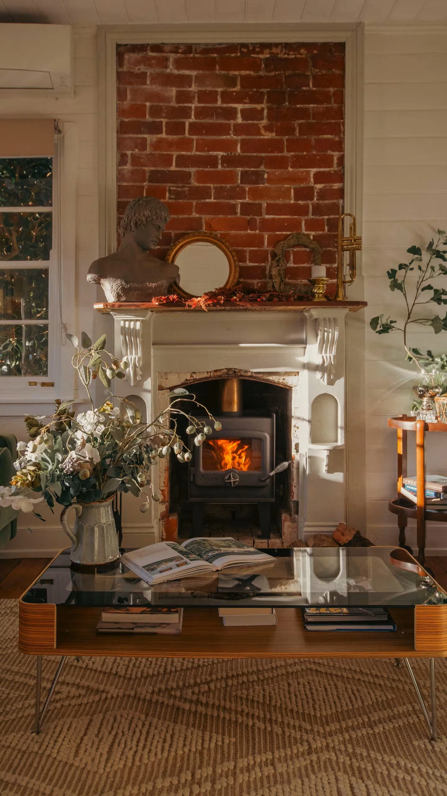 Living room with a fireplace, bust sculpture, mirror, candles, plants, coffee table with books, book open, and a rug.
