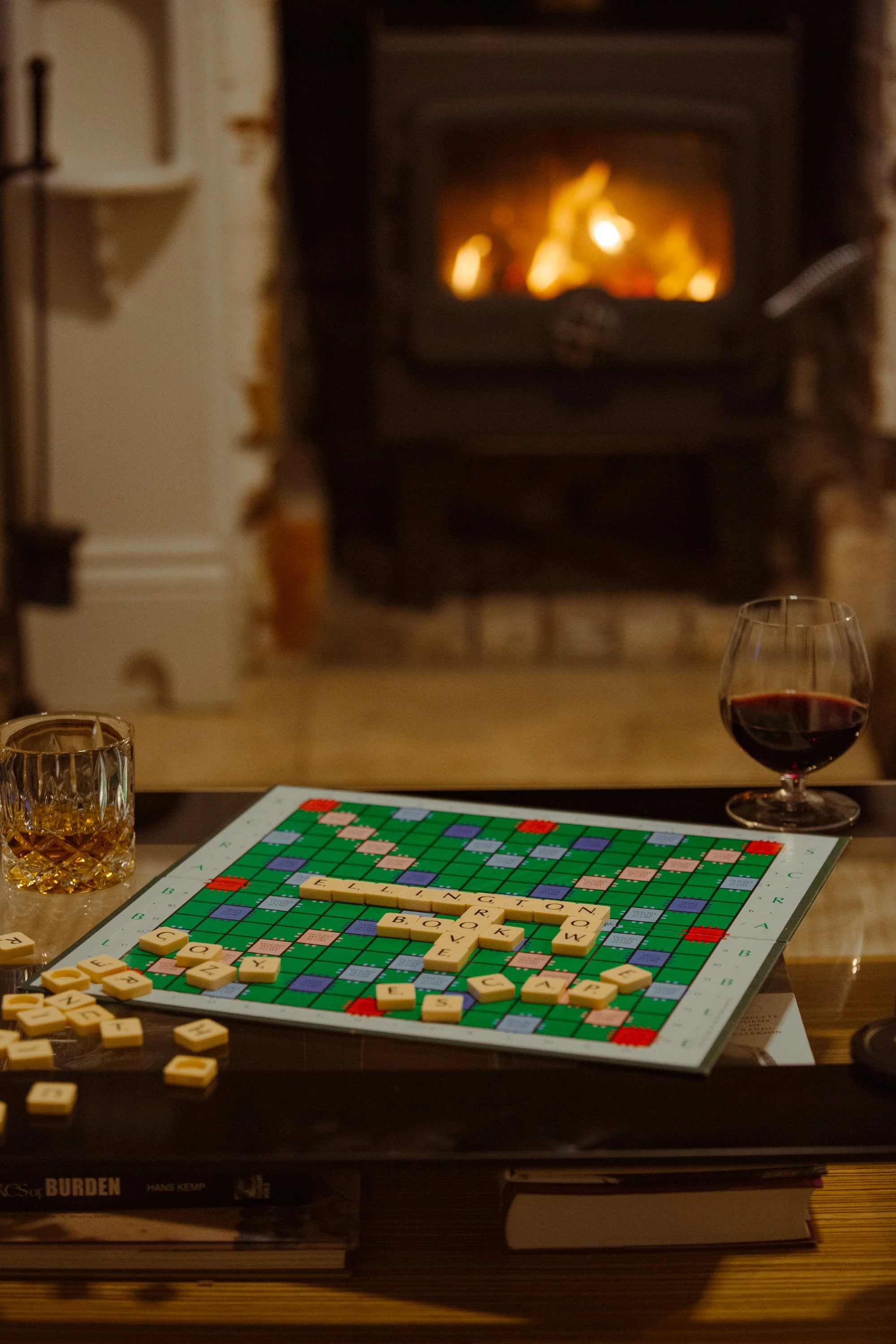 A Scrabble game in progress on a table with a fireplace in the background, flanked by a glass of whiskey and a glass of red wine, and some books underneath the table.