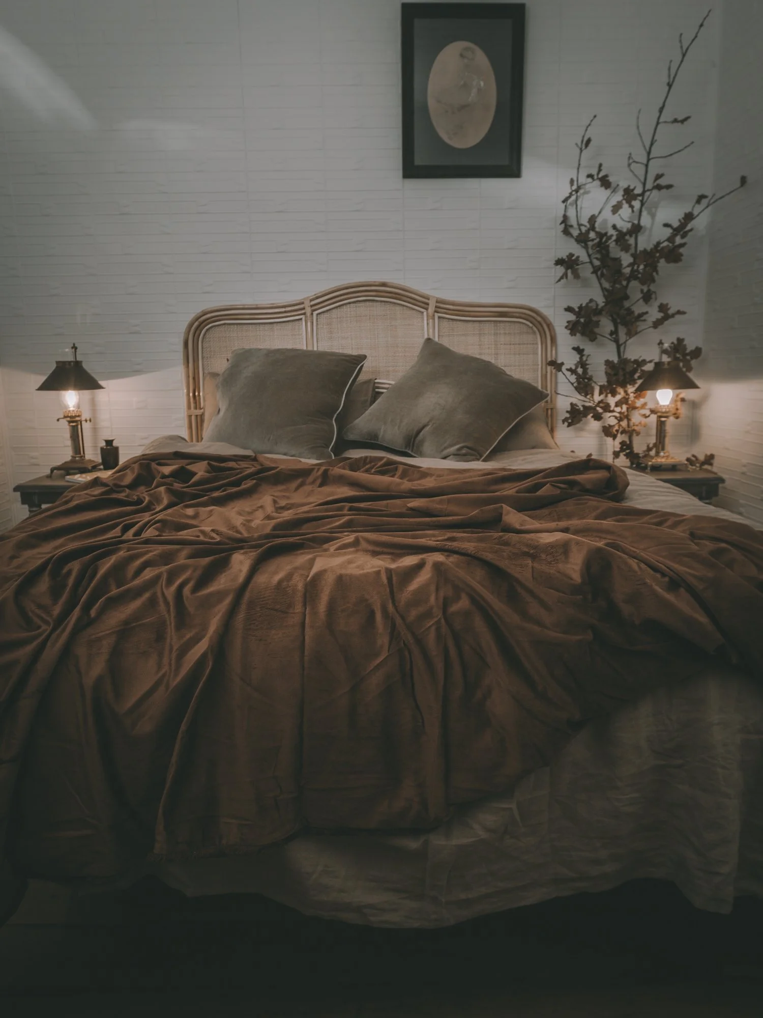 A cozy bedroom with a rattan headboard, beige and brown bedding, two large green pillows, matching lamps on nightstands, a framed moon illustration above the bed, and a decorative tree branch with leaves in the corner.