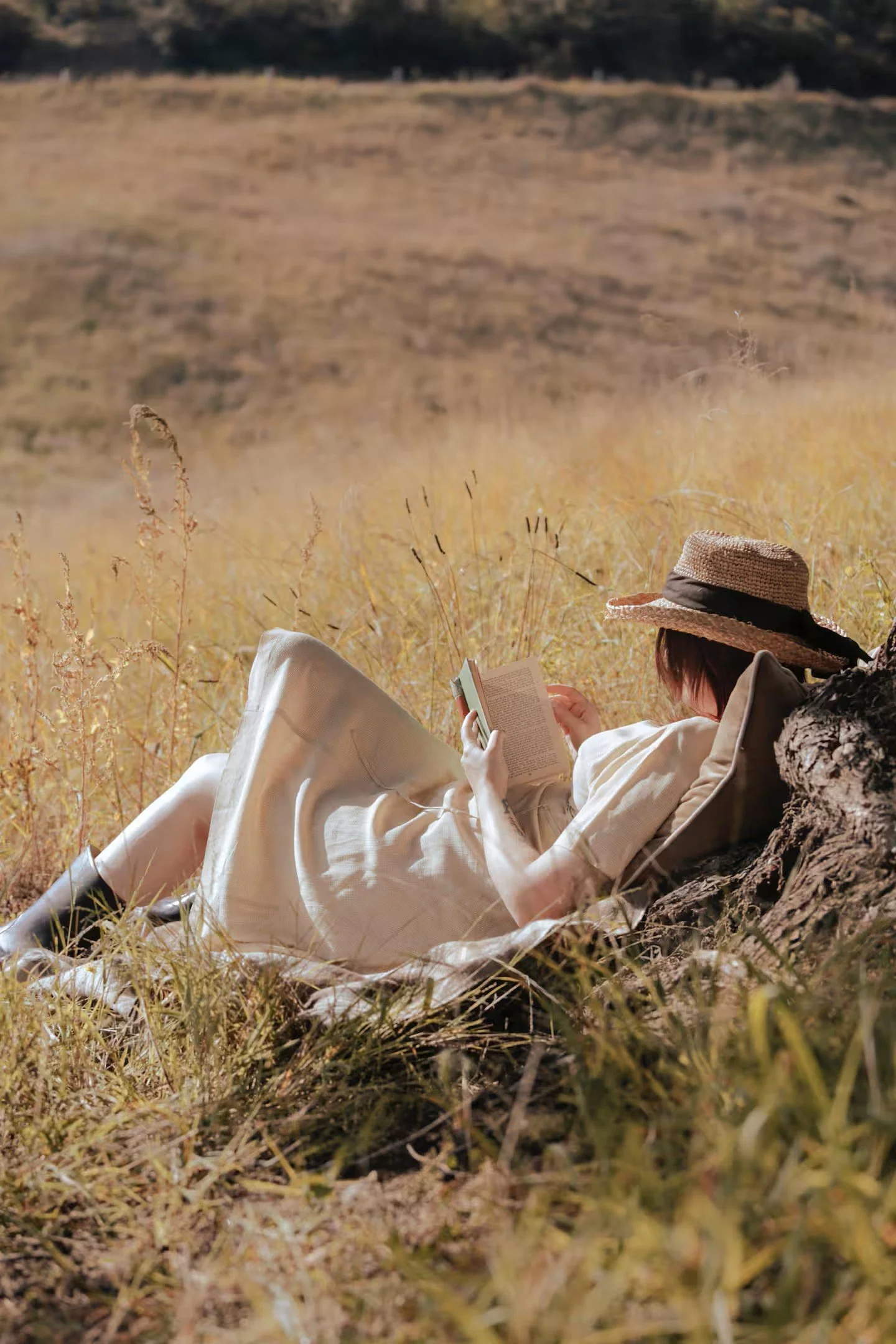 A woman in a white dress and straw hat lying on the grass, reading a book in a field during daytime.