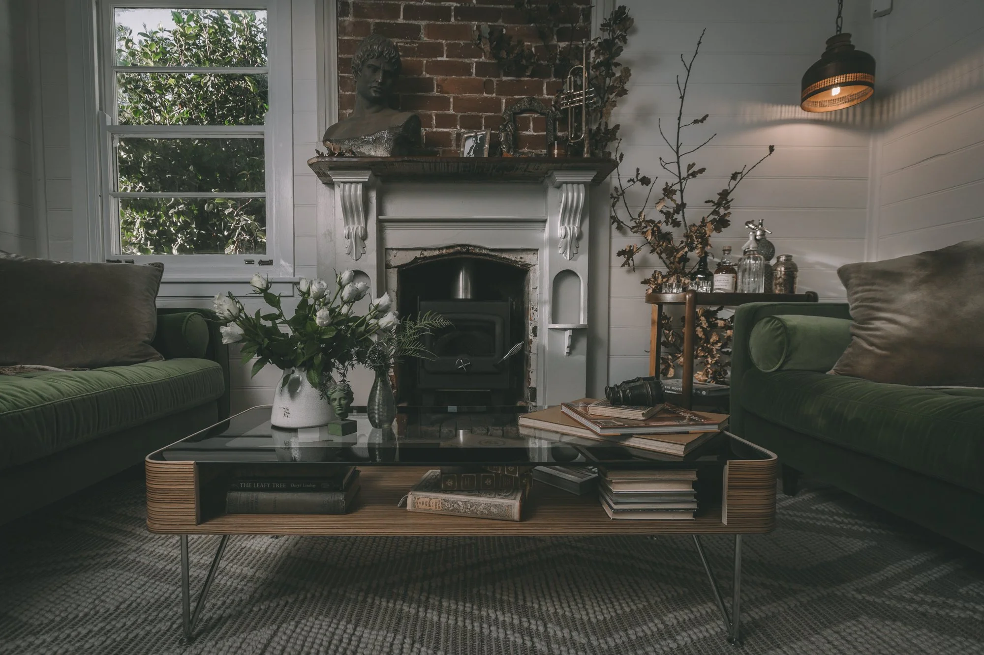 Cozy living room with a white fireplace, green sofas, a glass coffee table with books and a flower vase, and a large window with greenery outside.