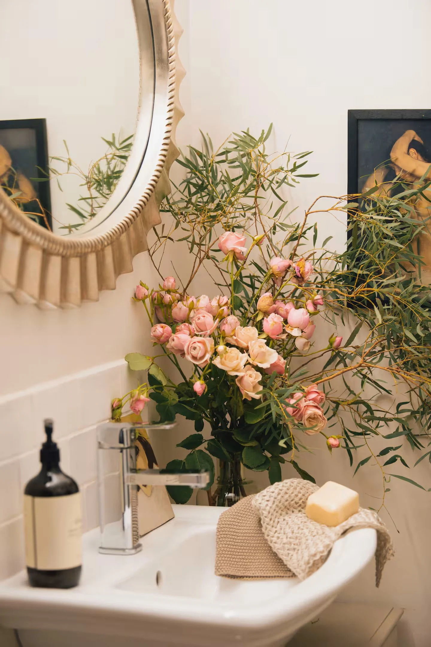 A bathroom sink with a beige towel and soap on it, a black soap dispenser, a vase filled with pink roses, and green branches, with framed artwork and a mirror on the wall behind.