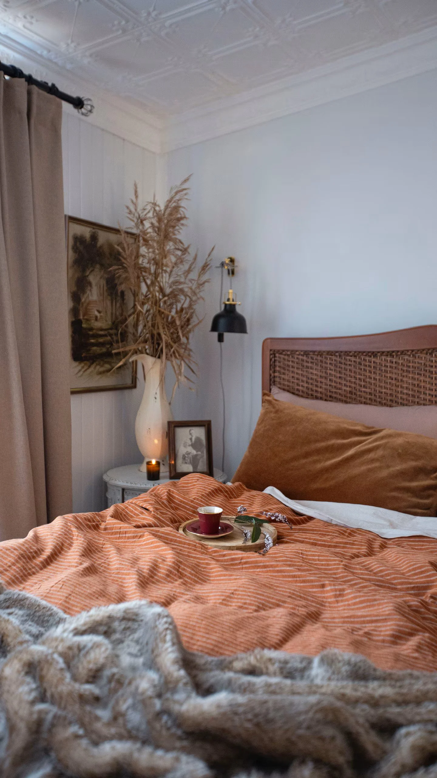 A cozy bedroom with a bed covered in an orange striped blanket and brown pillows. On the bed, there is a tray with a cup of tea and decorative sprigs. To the side, a nightstand holds a vase with dried plants, framed photographs, and a candle. The wal