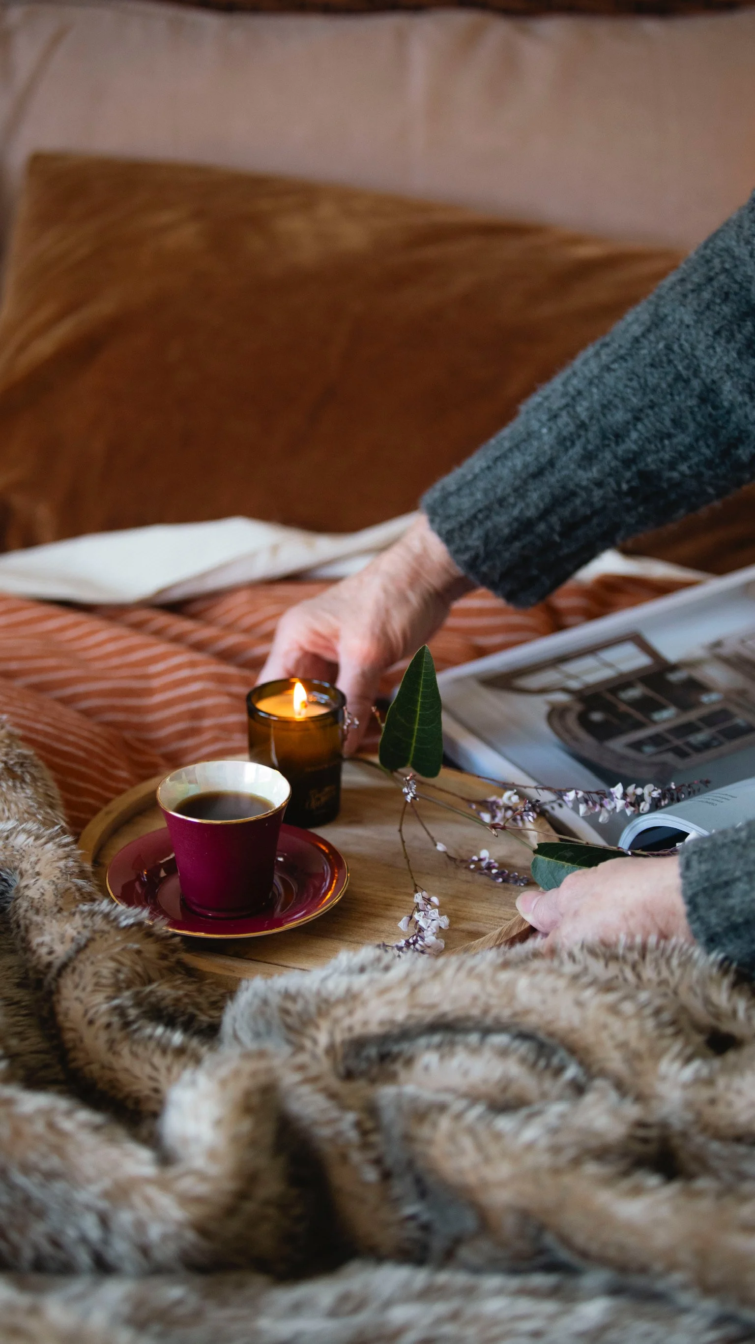 An elderly person's hands arranging a tray with a lit candle, a cup of coffee on a saucer, a branch with leaves and flowers, placed on a bed with a blanket, magazine, and pillows in the background.