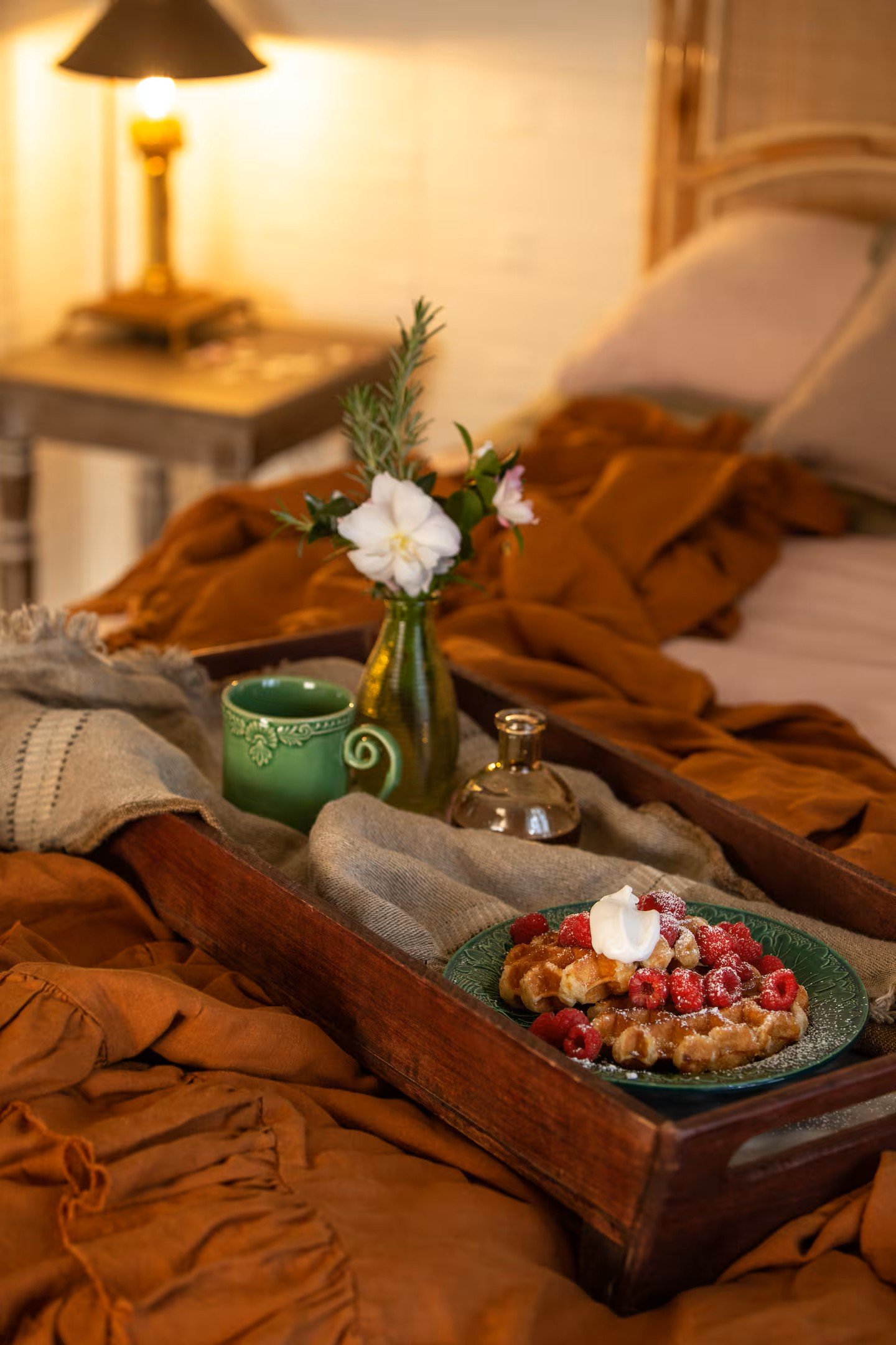 A bed with brown and beige blankets, a tray with waffles topped with strawberries and whipped cream, a green mug, a small glass bottle, and a small flower vase with white flowers on it, in a cozy bedroom with warm lighting.