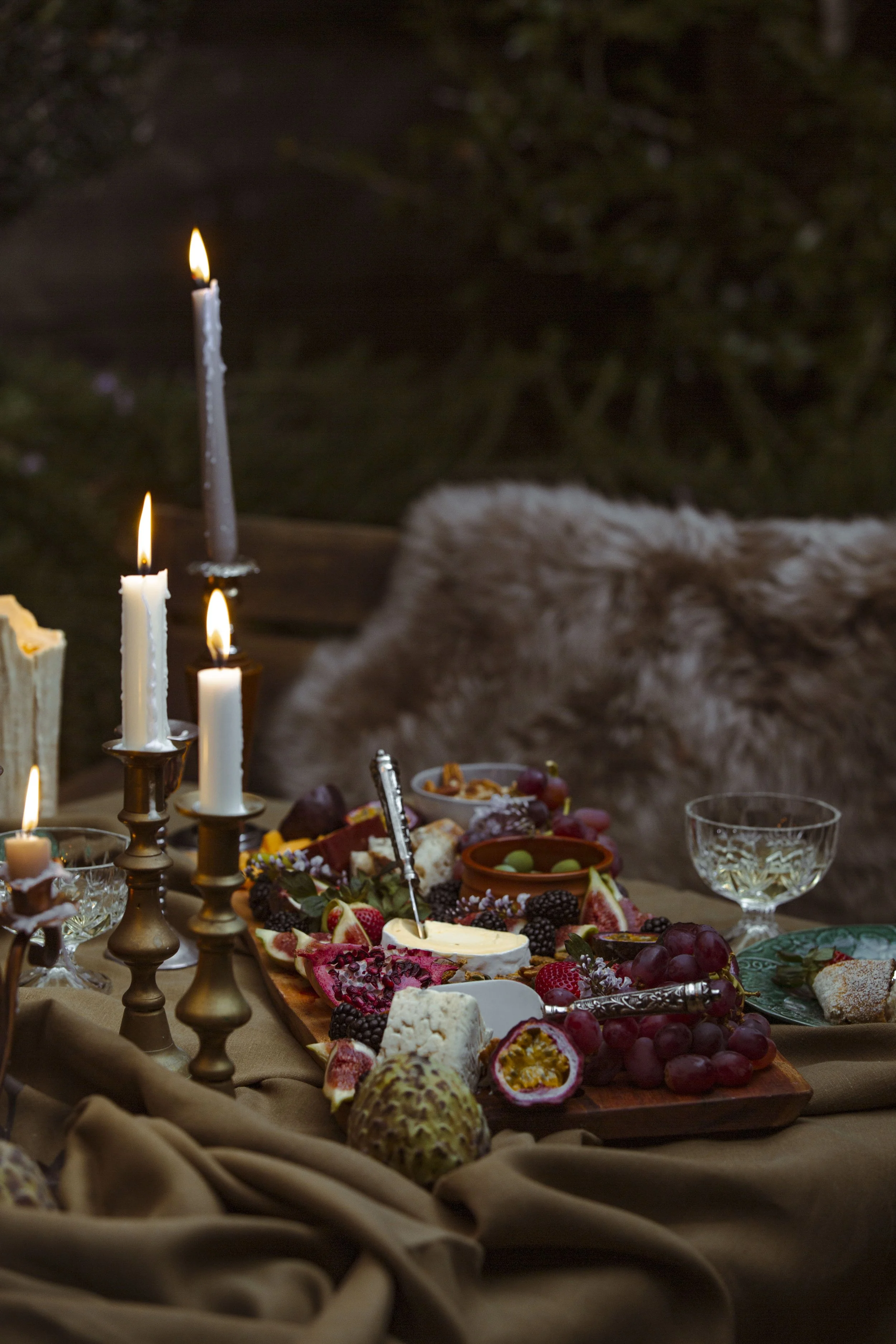 A table decorated for a celebration with lit candles, a wooden tray with assorted cheeses, fruits like grapes and passion fruit, and various snacks, surrounded by elegant glassware and beige fabric, with a furry blanket in the background.