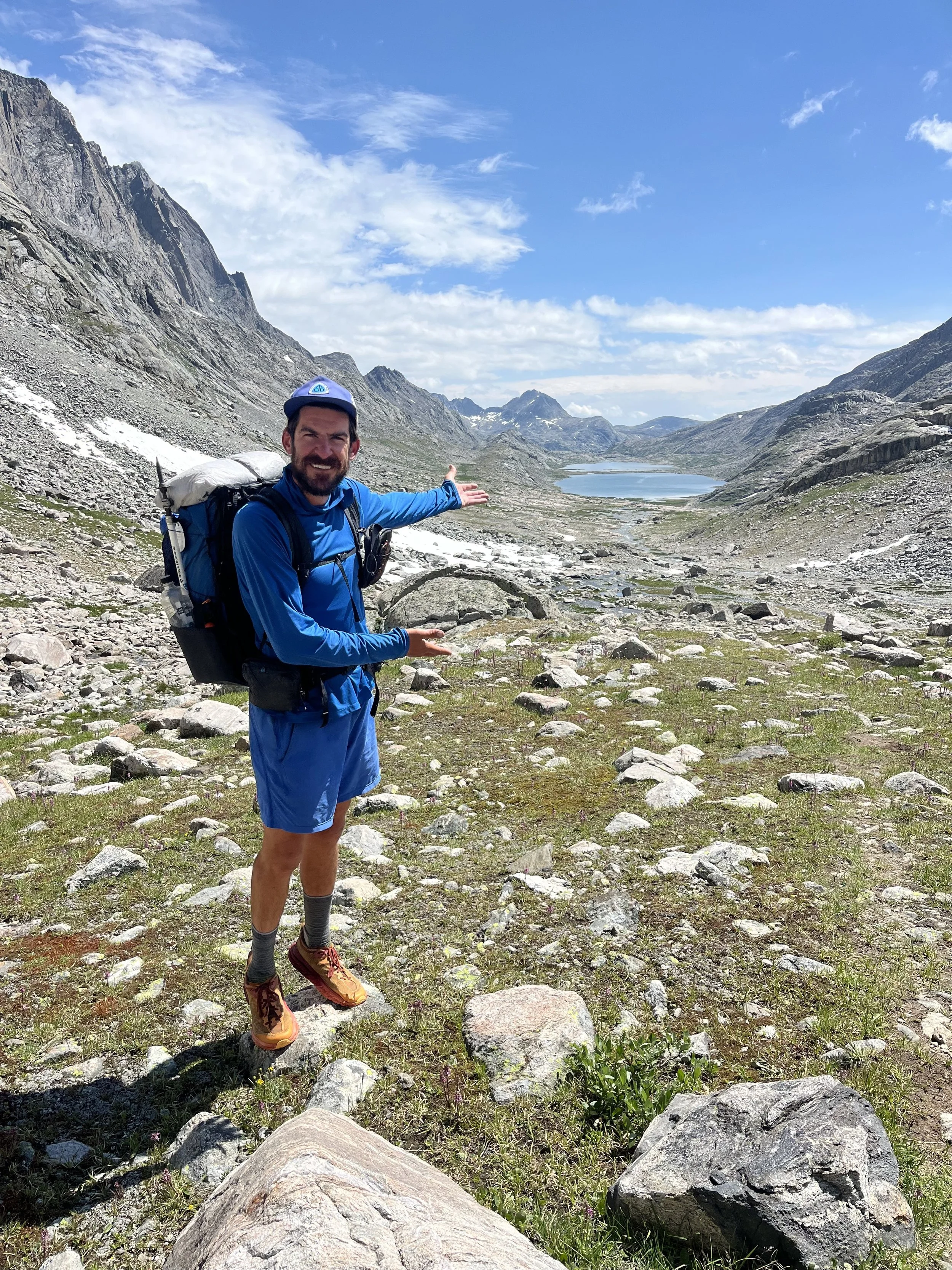 Cameron Virjee hiking s in a mountainous landscape pointing to a lake in the distance.