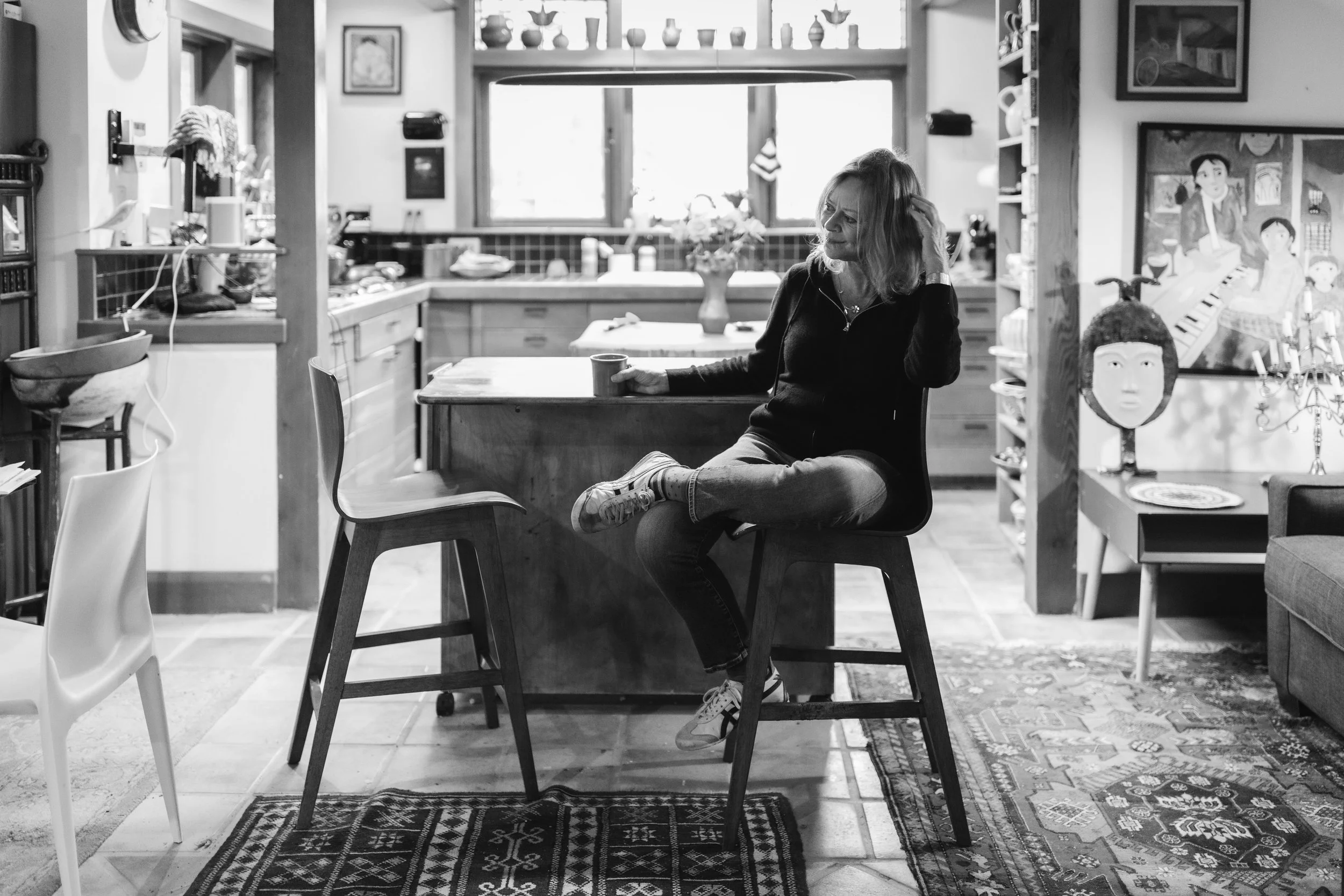 A woman sitting on a high chair with one leg crossed over the other, holding a mug, in a cozy kitchen with eclectic decor.