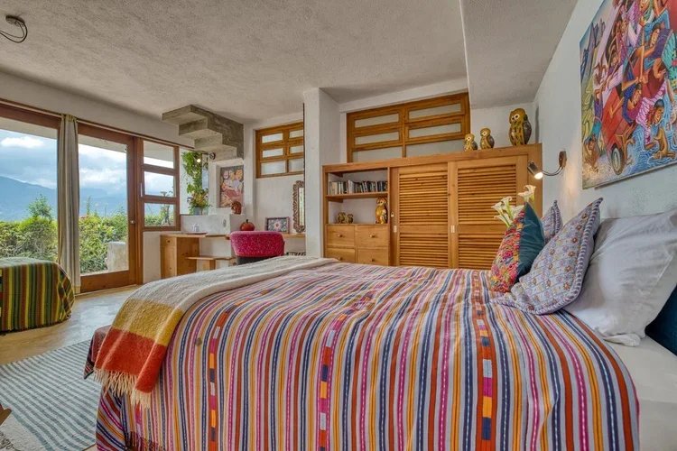 A colorful bedroom with a bed that has a striped multicolored bedspread, throw pillows, and a blanket. There are wooden cabinets and shelves, artwork on the walls, large windows with curtains, and a view of greenery outside.