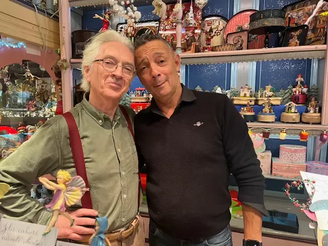 Two men stand inside a store filled with holiday decorations and collectibles. One man wears a green shirt with suspenders and glasses, holding a small fairy figurine. The other man wears a black sweater. Both are smiling and posing for the photo.