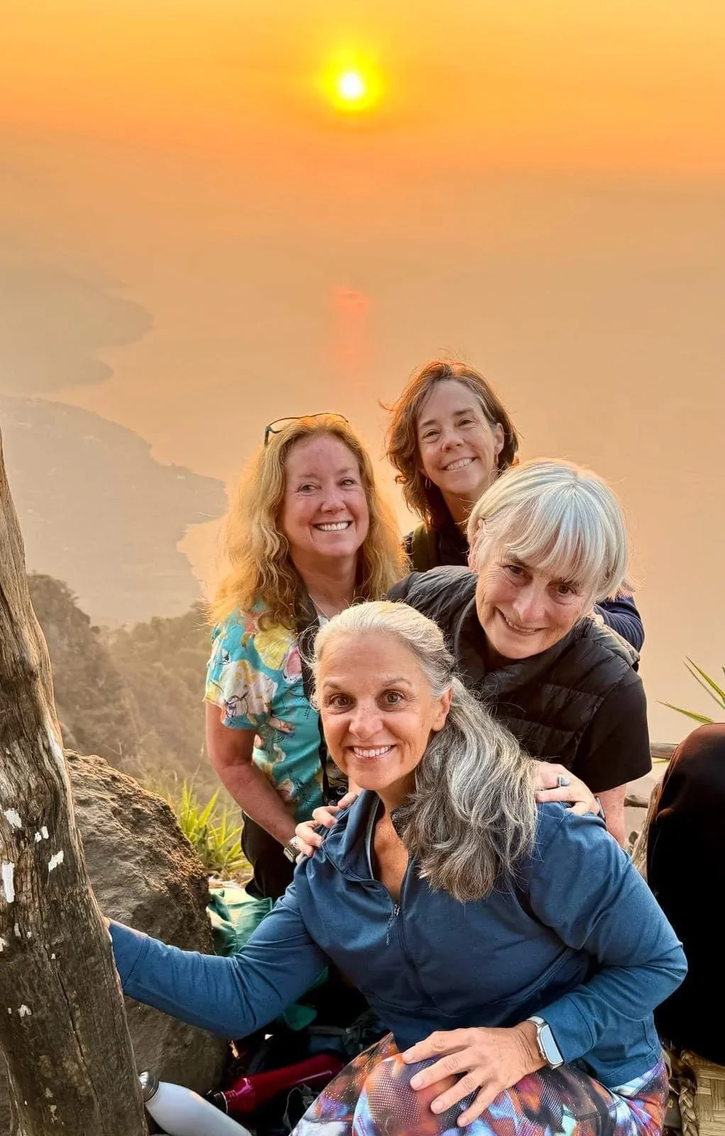Five women smiling together outdoors at sunset, with an orange sky and body of water in the background.
