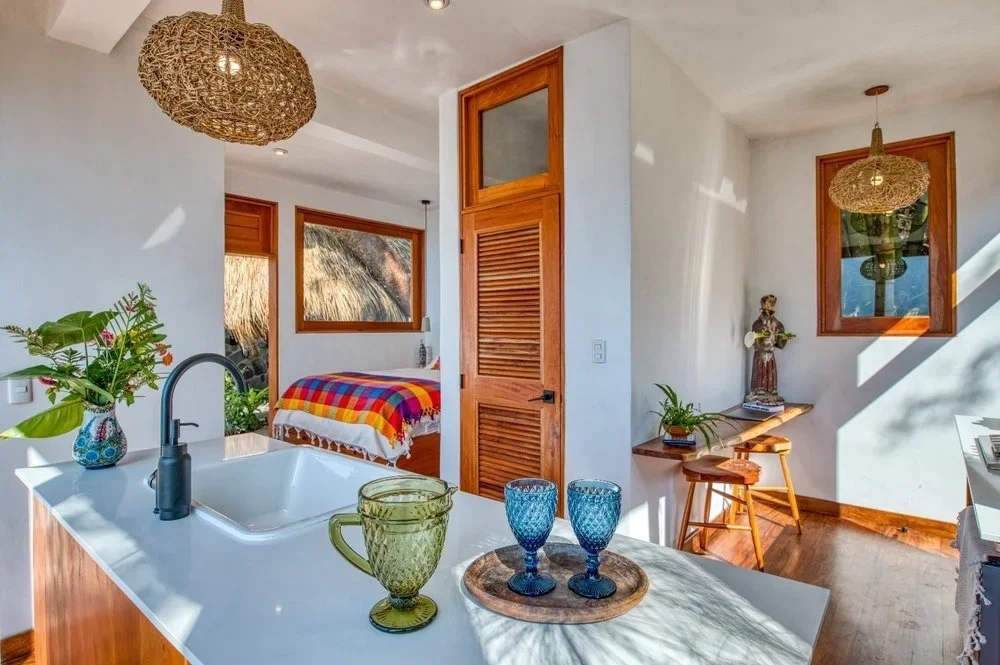 A cozy interior space showing a kitchen with a sink, a vase with flowers, and two drinking glasses on a wooden tray. Behind the kitchen, there is a bedroom with a bed covered in a vibrant, multicolored blanket. The room features wooden trim around the windows and doors, and decorative woven light fixtures hanging from the ceiling.