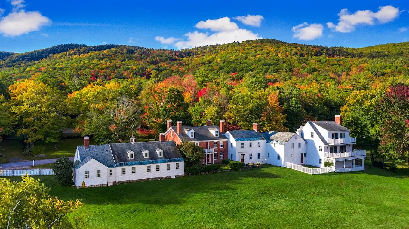 A group of white and red houses with spacious green lawns in front of a densely forested hillside during fall, with clear blue skies and a few clouds.