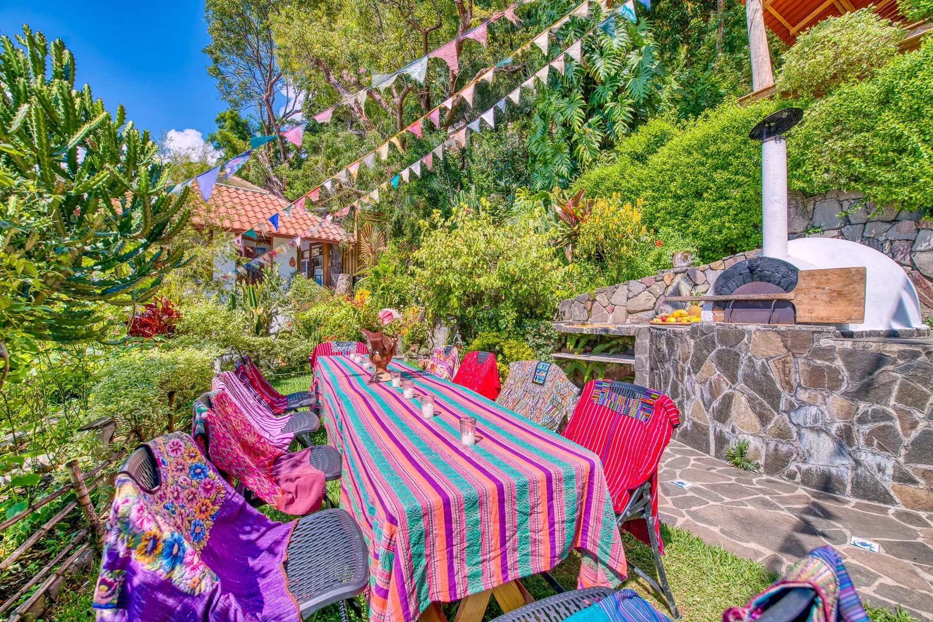 Outdoor garden gathering with a long table covered in a colorful striped tablecloth, surrounded by chairs with vibrant fabrics, and decorated with flags hanging overhead. There is a stone pizza oven in the background, lush green plants and trees all around, and a small house with a red-tiled roof partially visible.