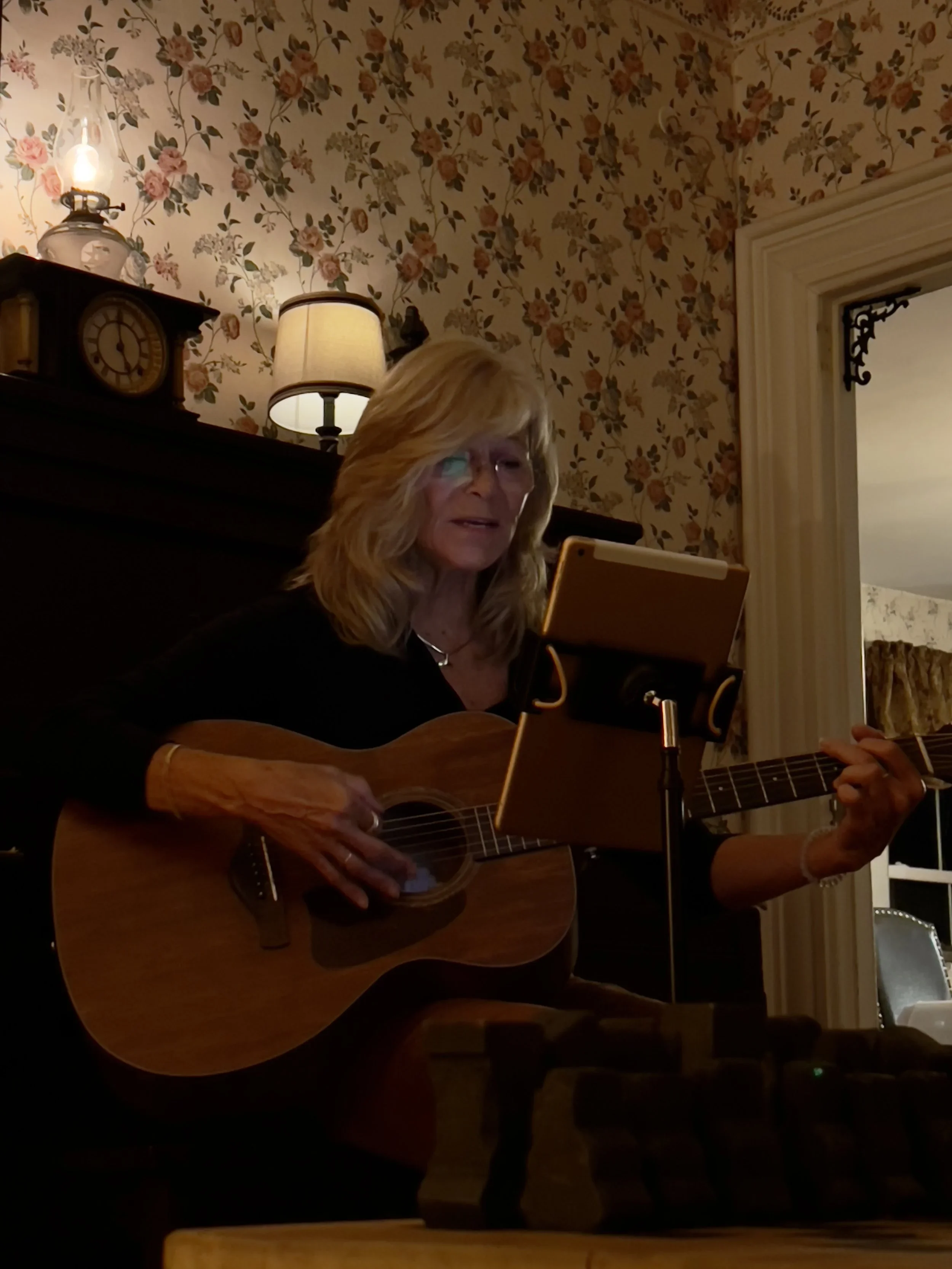 A woman wearing glasses playing an acoustic guitar indoors in a room with vintage floral wallpaper, a clock, and lamps on the fireplace mantel.