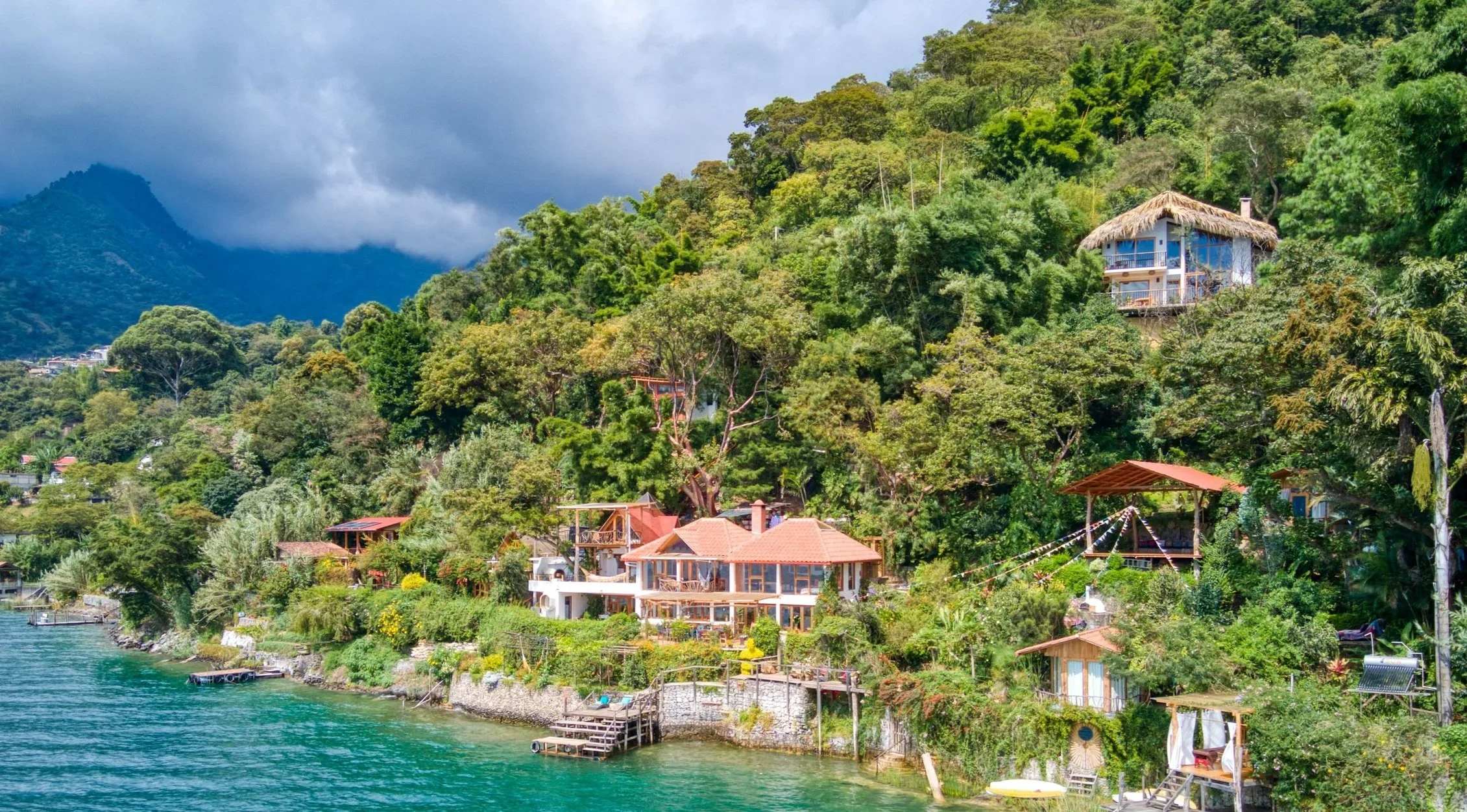 Houses and villas on a hillside by a lake surrounded by lush green trees and mountain scenery under cloudy sky.