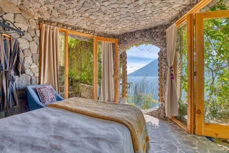 A cozy bedroom with stone walls and a large window showing a mountain view, featuring a bed with a white blanket and a yellow throw, a blue armchair, and curtains.