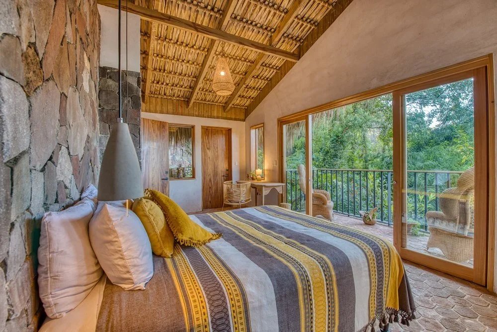 Bedroom with a bed covered in striped yellow, gray, and brown blankets, white and yellow pillows, stone accent wall, sloped wooden ceiling, large glass sliding door leading to a balcony with wicker chairs, and lush greenery outside.