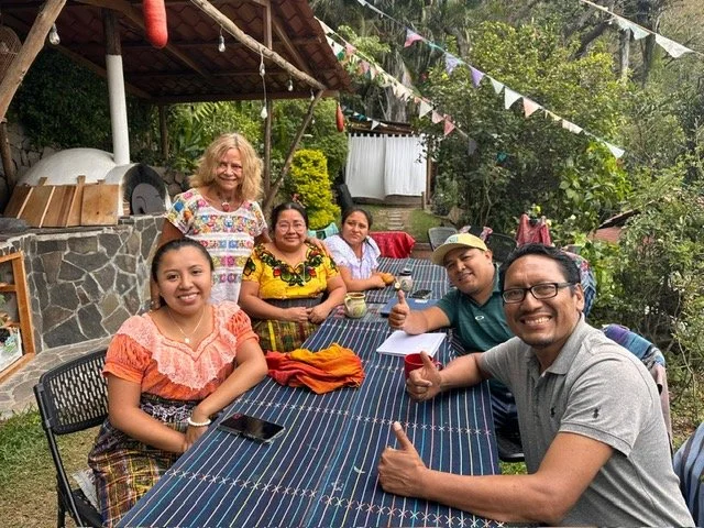 A group of six people sitting and standing around a long striped table outdoors, enjoying drinks and smiling, with colorful flags hanging above and a lush garden in the background.
