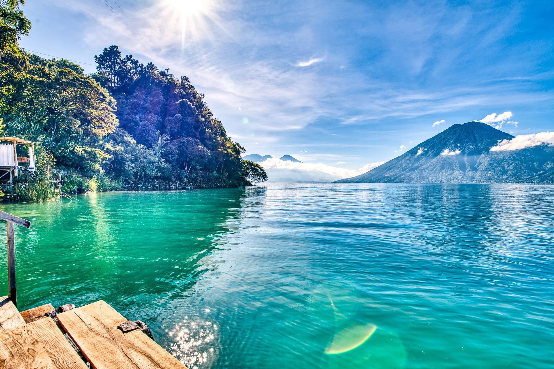 Scenic view of a lake with turquoise water, surrounded by lush green mountains and dotted with volcanoes in the background under a bright blue sky with wispy clouds.