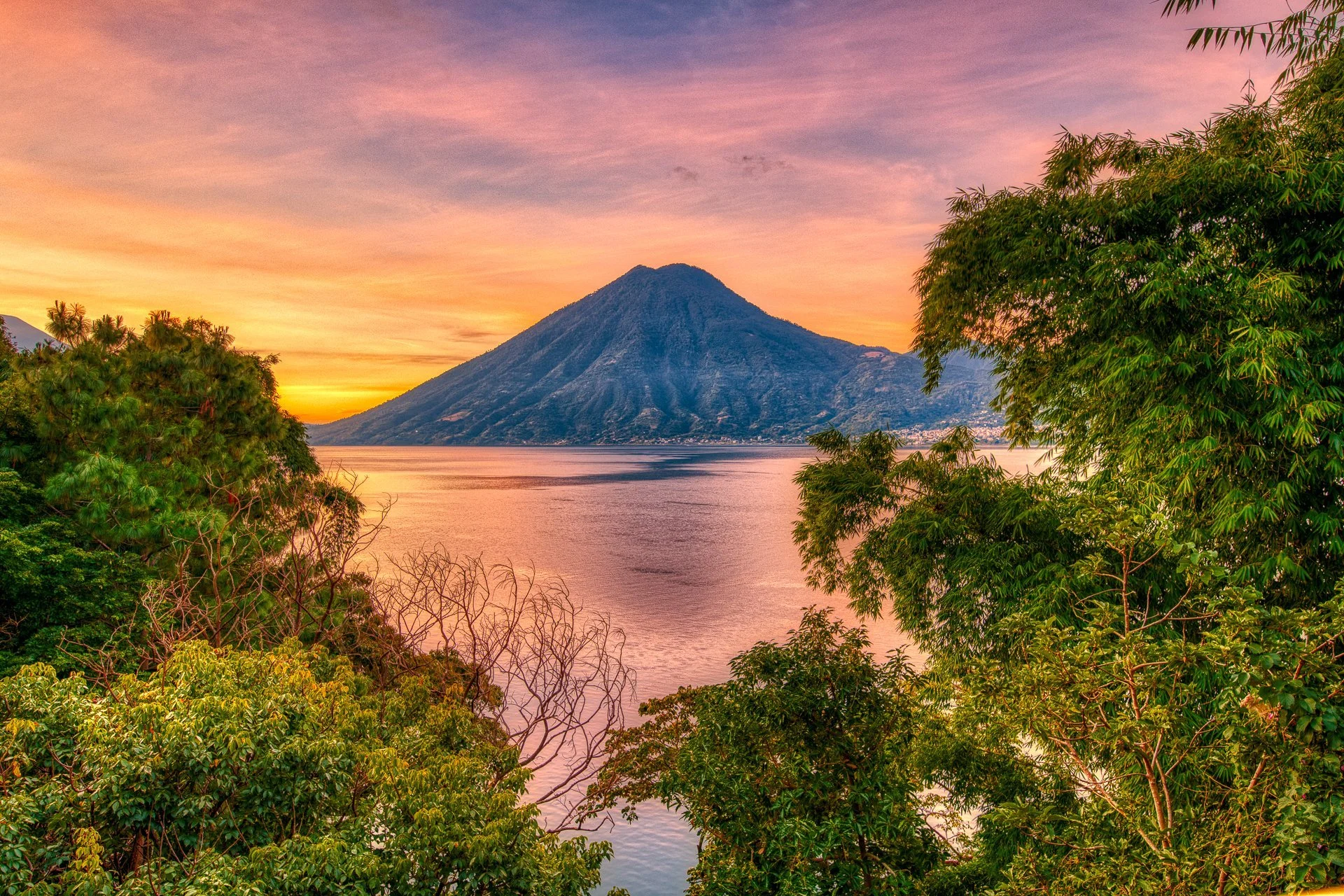 A scenic view of a volcano near a body of water at sunset, framed with lush green trees.