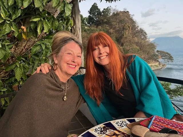 Two women smiling and sitting outdoors near a lake, with green trees and cloudy sky in the background.
