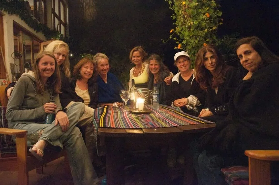 Group of nine women sitting around a table outdoors at night, smiling, with drinks and a candle on the table, in a cozy setting surrounded by greenery.