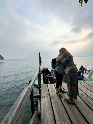 Two women hugging on a wooden pier by the water with boats docked nearby, under a cloudy sky.