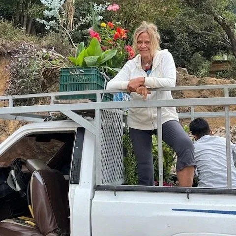 A woman standing in the back of a white pickup truck with a metal railing, surrounded by plants and flowers, with one person kneeling beside the truck.