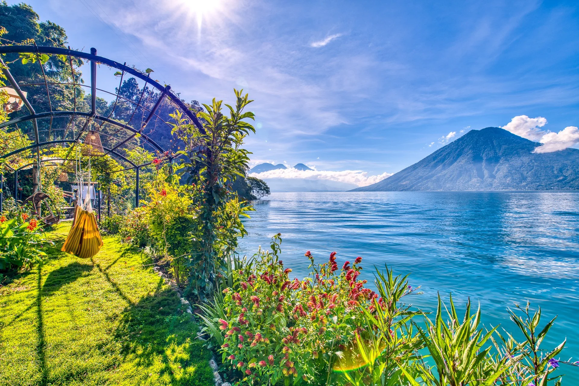 A lush garden next to a large blue lake with a mountain in the background under a bright sky.
