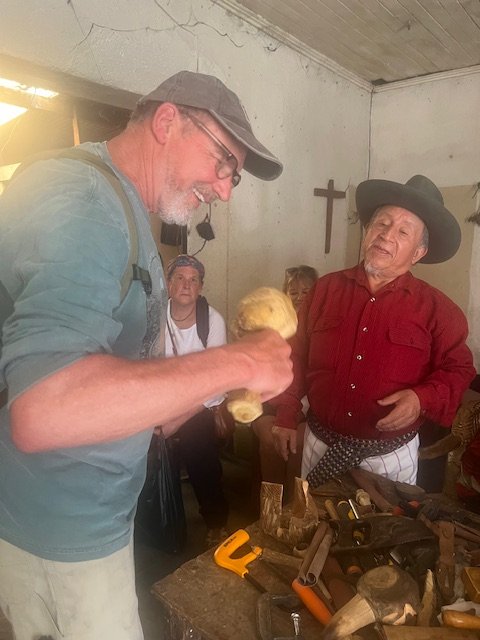 A man in a blue shirt and gray cap showing a large, yellowish mushroom to a woman in a red shirt and black hat in a rustic room with wartime artifacts.