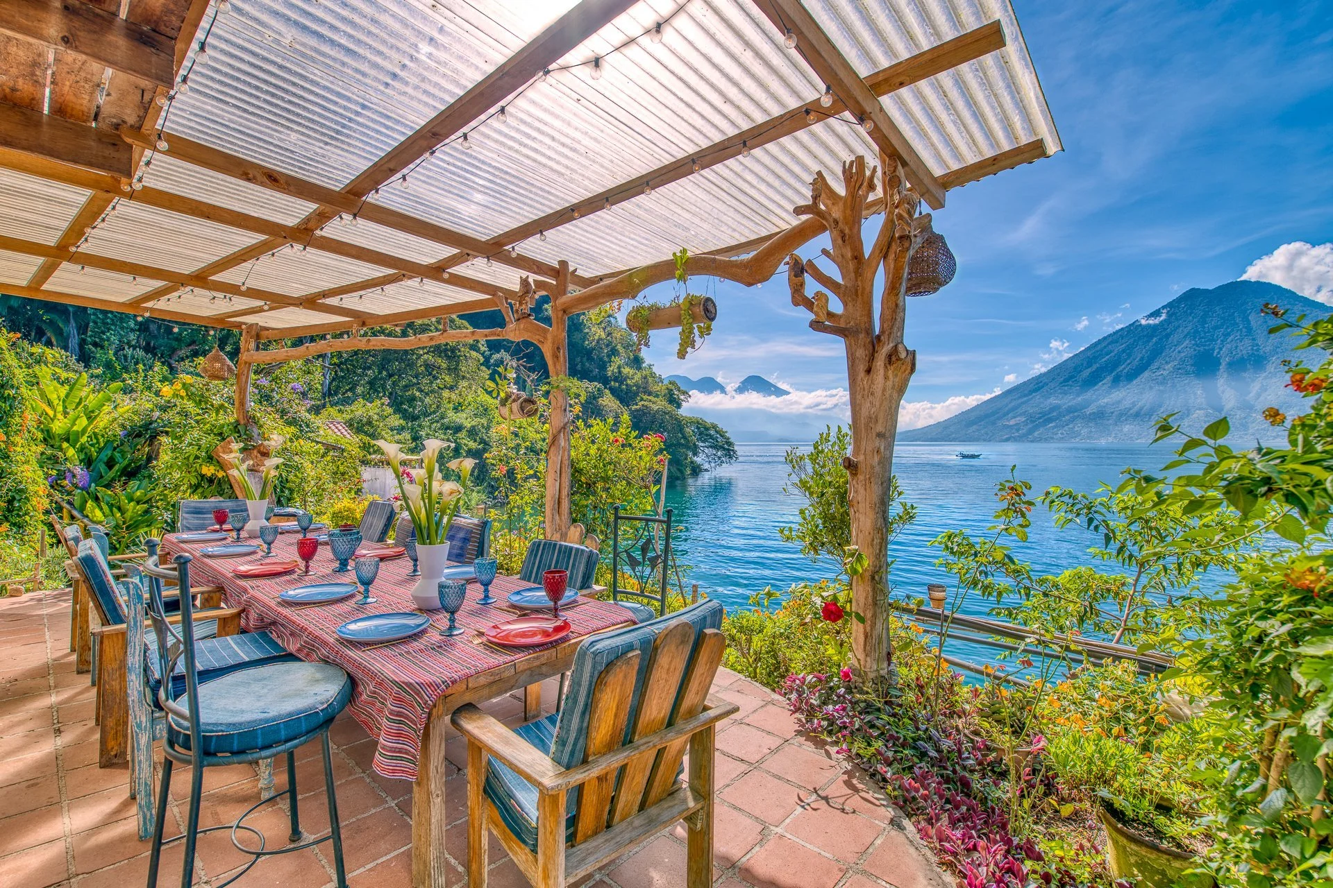 Outdoor dining area with a long table set with colorful plates and glasses, under a wooden and corrugated metal roof, overlooking a lake with lush greenery and mountains in the background.