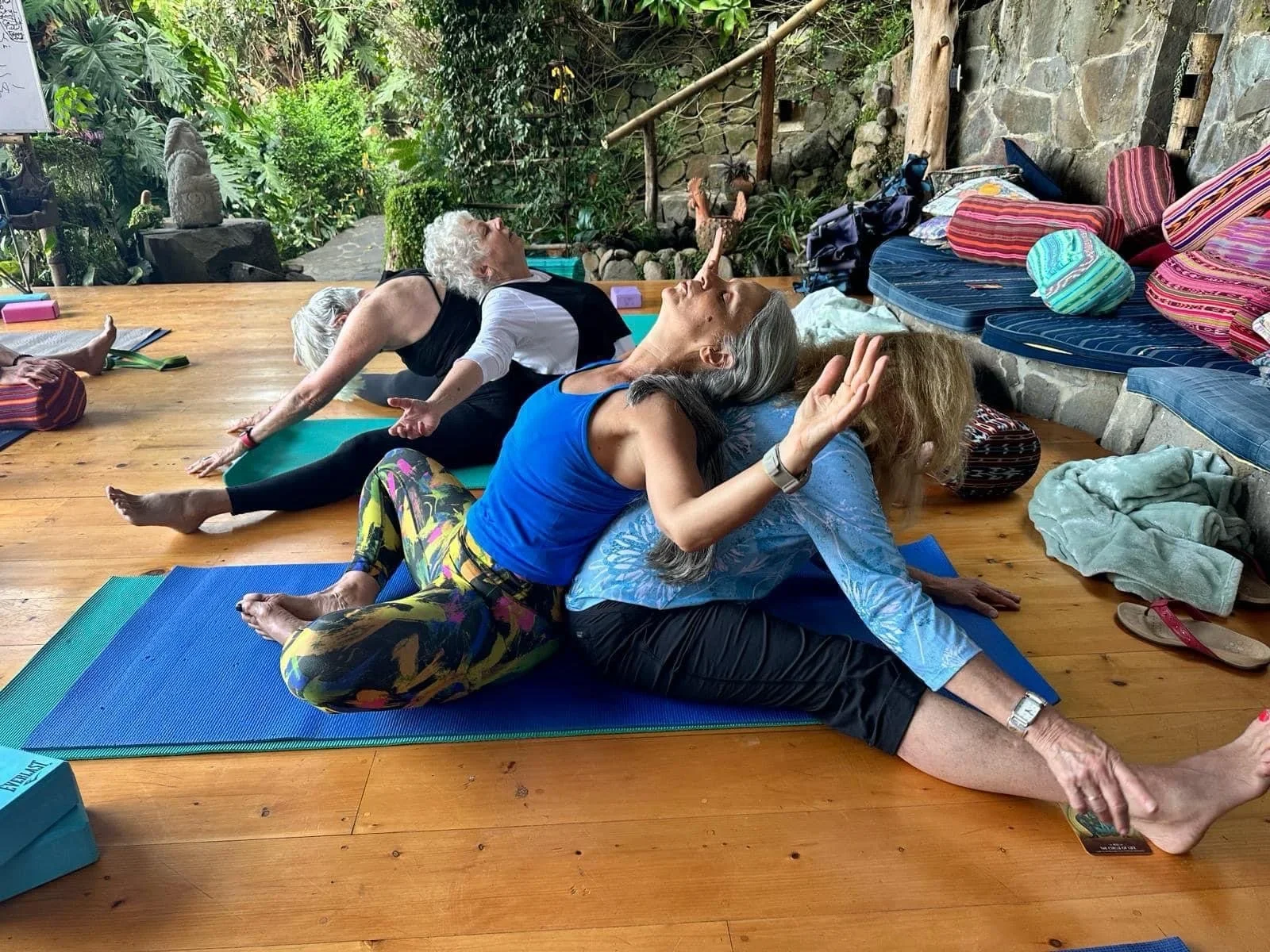 Four women practicing yoga on mats in a tranquil, outdoor, wooded space with stone and wooden elements.