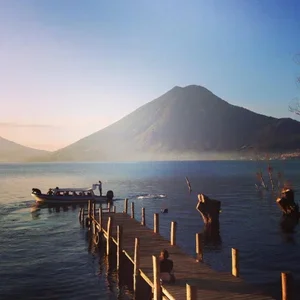 A wooden pier extends into a body of water with a boat nearby, set against a large mountain landscape at sunset or sunrise.