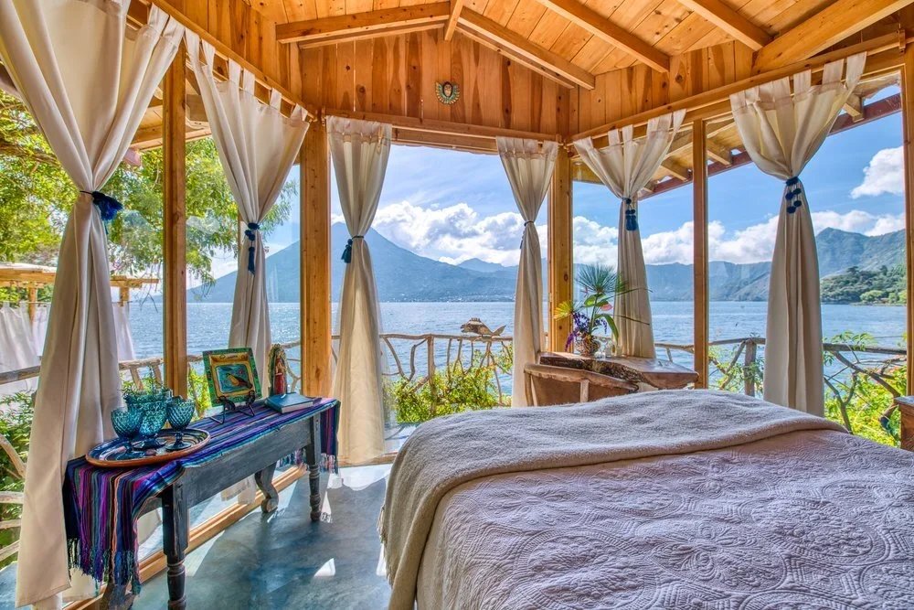 Bedroom with large windows and curtains overlooking a lake and mountains.